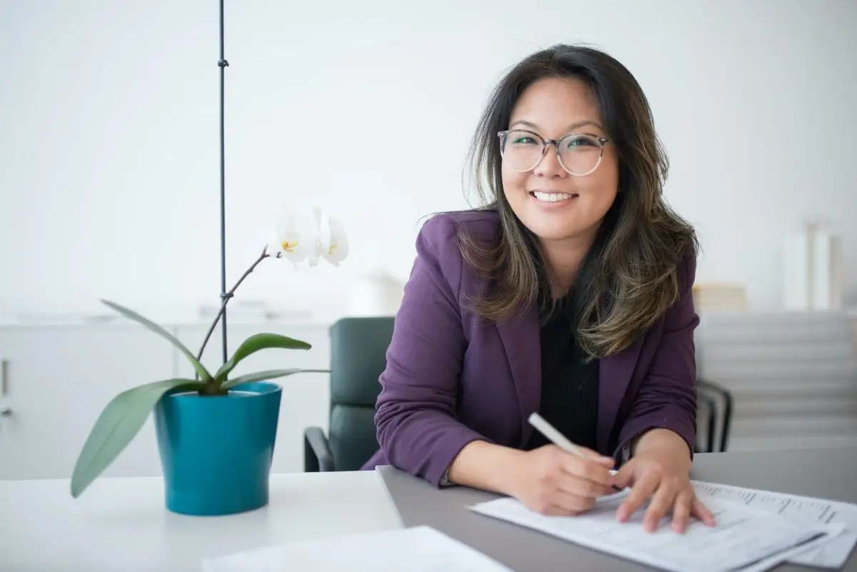 A smiling woman with glasses sits at a desk, writing on papers with an orchid and a blue pot on the desk.