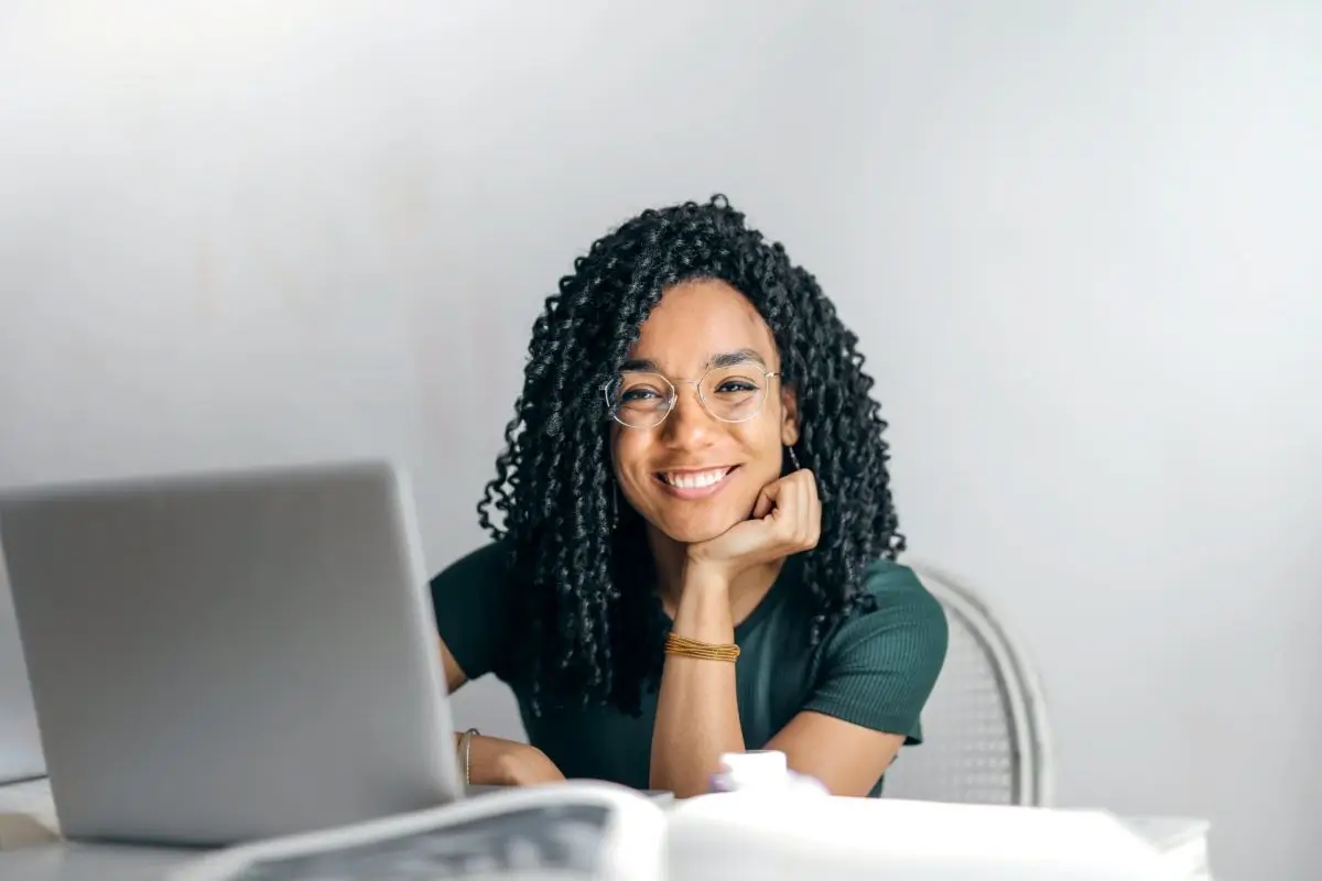 A woman with curly hair and glasses smiling at the camera, sitting at a desk with a laptop and papers in front of her
