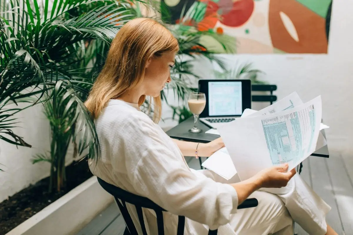 A woman reviews documents at a workspace with a laptop and a drink on the table, surrounded by a vibrant artwork backdrop.