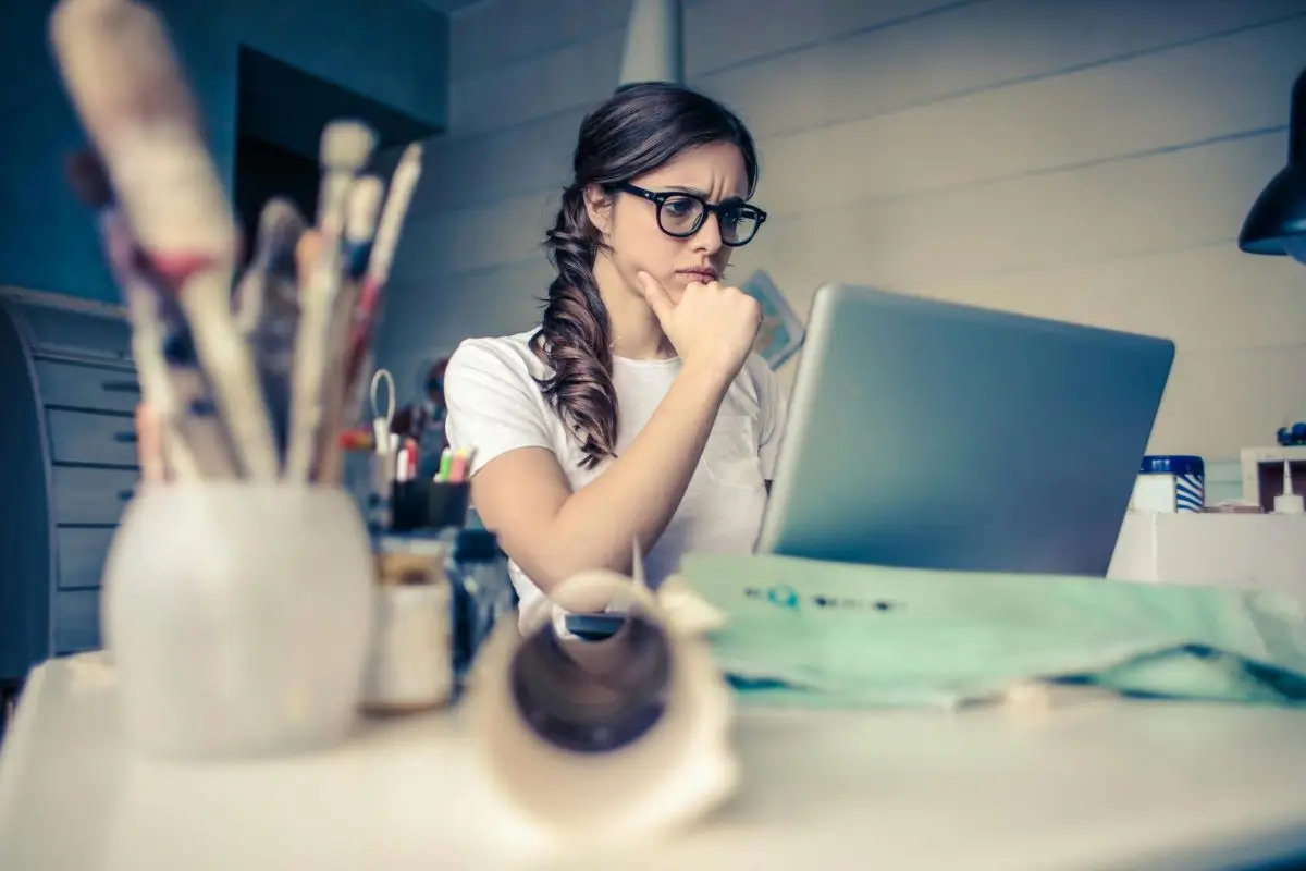 A woman with glasses and a braid looks pensively at her laptop in a cluttered office space