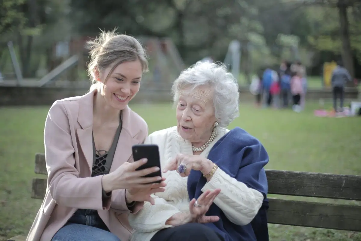 A young woman and an elderly woman sitting on a park bench, looking at a smartphone together with a joyful expression